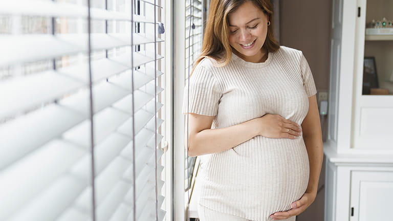 Zwangere vrouw bij het raam met handen op buik